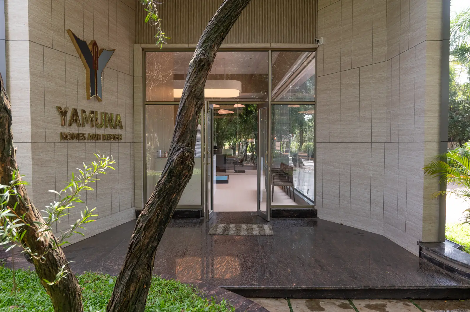 The entrance to the same showroom, a glazed door set into stone cladding above a polished granite threshold, with brass lettering reading Yamuna Homes and Design on the wall alongside